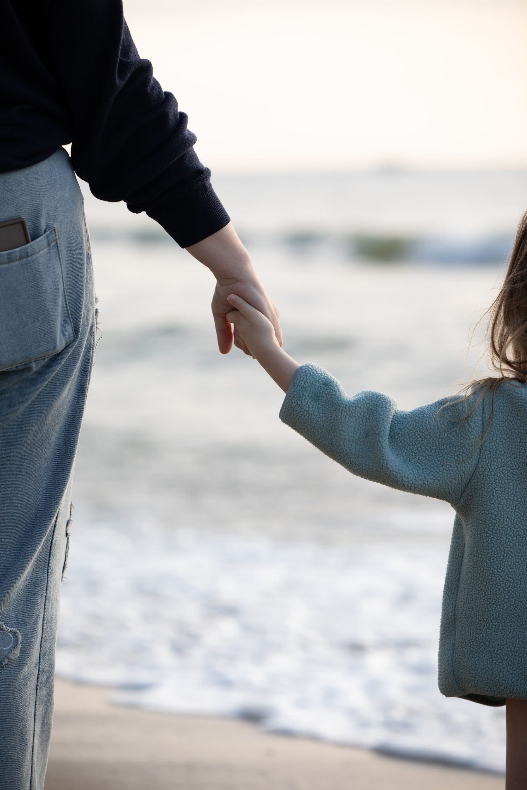 An adult hand holding a small child’s hand as they walk beside the water.